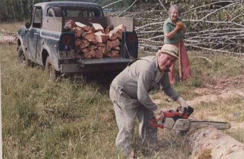 Man using a chainsaw to cut wood with a truck loaded with firewood in the background and a woman doing yardwork