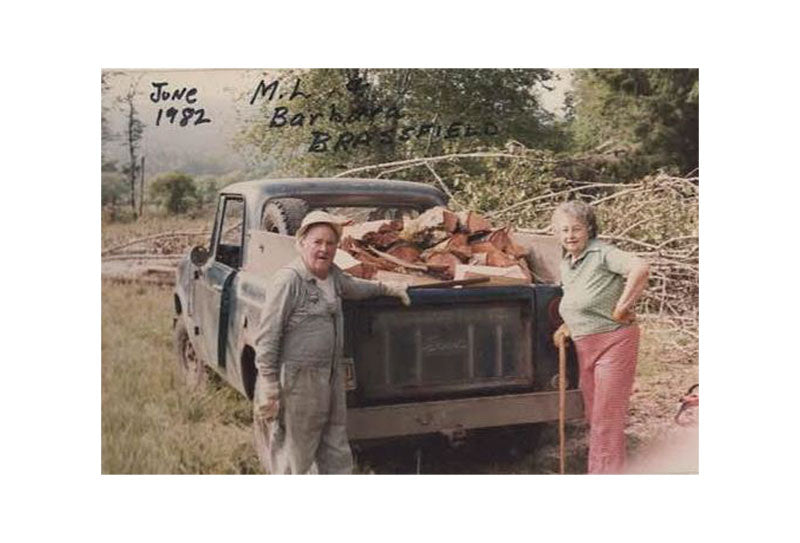 Two people standing next to a pickup truck filled with wood, with text on the image indicating a date and location.